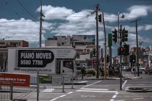 A busy urban street corner with a truck labeled 'Piano Moves' parked near pedestrian crossing signs. Several people stand at the crosswalk, waiting to cross. Overhead, power lines intersect the cloudy sky. In the background, a variety of shops and a billboard advertising an internet bundle are visible.