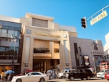 A lively urban scene with the iconic Dolby Theatre as the central focus. The building features modern architecture with large glass windows, beige concrete, and a prominent marquee sign. The street is bustling with vehicles, including a white sedan and a black SUV, and there are people gathered on the sidewalk under colorful umbrellas. Nearby, signs for shops such as Sephora are visible, and a pedestrian crossing sign is in the foreground.