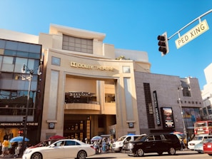 A lively urban scene with the iconic Dolby Theatre as the central focus. The building features modern architecture with large glass windows, beige concrete, and a prominent marquee sign. The street is bustling with vehicles, including a white sedan and a black SUV, and there are people gathered on the sidewalk under colorful umbrellas. Nearby, signs for shops such as Sephora are visible, and a pedestrian crossing sign is in the foreground.