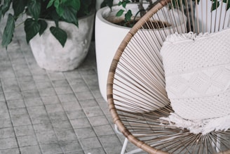 A stylish chair with a woven design sits on a tiled floor, featuring a textured white cushion. Surrounding the chair are potted plants with green leaves, adding a touch of nature to the scene.