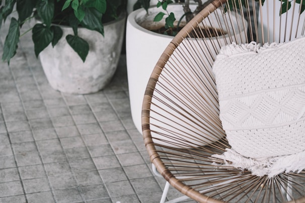 A stylish chair with a woven design sits on a tiled floor, featuring a textured white cushion. Surrounding the chair are potted plants with green leaves, adding a touch of nature to the scene.