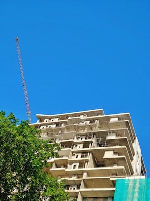 A partially constructed multi-story building with exposed concrete structures and scaffolding. A tall construction crane is visible on the left. Lush green trees are present in the foreground under a clear blue sky.