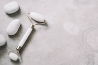 A jade roller lies on a smooth, light gray surface surrounded by smooth white stones. The image conveys a sense of calm and relaxation, often associated with skincare routines.