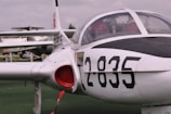An aircraft parked on a green surface with numbers 2835 prominently displayed on its side. The cockpit is visible with a clear canopy, and part of another aircraft is seen in the background with similar livery.