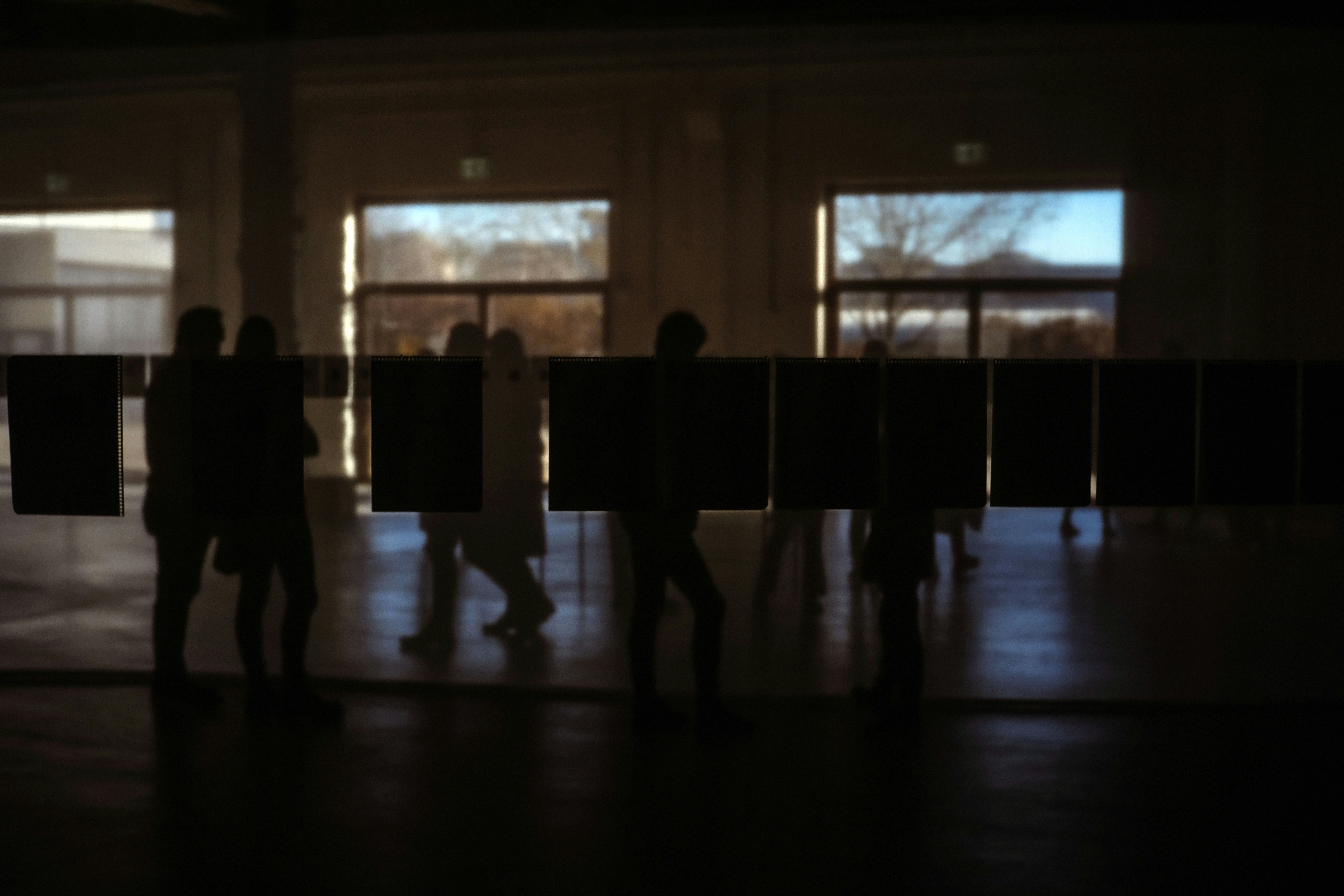 Gallery installation with hundreds of silk panels hanging from high ceiling, warm amber and cream tones, visitors silhouettes below, dramatic lighting