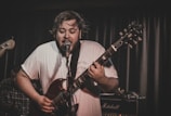 A musician passionately performs on stage, playing a guitar and singing into a microphone. The setting appears to be a dimly lit concert venue with visible stage equipment, including a Marshall amplifier in the background. The musician is wearing a white t-shirt and conveys a sense of intense emotion and engagement.