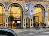 A large, ornate storefront with arched windows showcases an interior filled with people. The exterior features intricate designs and a prominent flag with an Apple logo. Bicycles are parked nearby, and pedestrians walk along the pavement.
