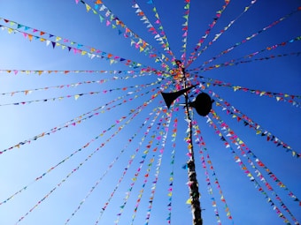Brightly colored triangular flags stretch from a central point on a tall pole against a clear blue sky. The flags are arranged in strings, fanning outwards in an orderly pattern. A black silhouette of a loudspeaker is visible near the top of the pole.