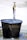 A brass faucet is turned on, allowing water to flow into a large, dark metal bucket placed on the ground. The bucket is visibly old and weathered, with water splashing around its edges, creating small puddles on the surface below.