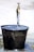 A brass faucet is turned on, allowing water to flow into a large, dark metal bucket placed on the ground. The bucket is visibly old and weathered, with water splashing around its edges, creating small puddles on the surface below.