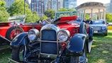 A collection of vintage and luxury cars displayed on a green lawn in an urban park setting. The foreground features a polished red and black classic car with prominent headlights and intricate detailing. Behind it, there are more vintage cars and a modern luxury vehicle. Towering city buildings and lush green trees surround the area, with a historical gazebo visible in the background.