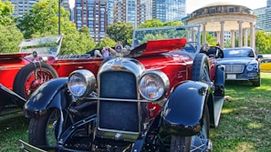 A collection of vintage and luxury cars displayed on a green lawn in an urban park setting. The foreground features a polished red and black classic car with prominent headlights and intricate detailing. Behind it, there are more vintage cars and a modern luxury vehicle. Towering city buildings and lush green trees surround the area, with a historical gazebo visible in the background.