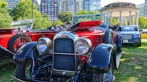 A collection of vintage and luxury cars displayed on a green lawn in an urban park setting. The foreground features a polished red and black classic car with prominent headlights and intricate detailing. Behind it, there are more vintage cars and a modern luxury vehicle. Towering city buildings and lush green trees surround the area, with a historical gazebo visible in the background.