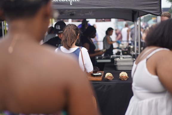 A group of people are gathered around a food vendor or stall at an outdoor event. Two burgers are displayed on a table beneath a canopy. The focus is on the back of a person with a bun hairstyle wearing a denim outfit. Others in the background are interacting with the vendor.