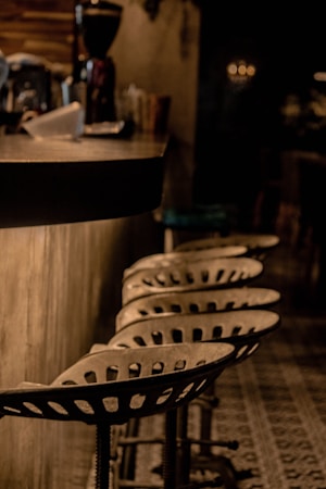 A dimly lit bar area features a row of industrial-style metal stools with perforated seats lined up against a counter. The counter is cluttered with various items including a paper cup and a large coffee grinder in the background. The overall ambiance is rustic and cozy.