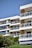 A modern apartment building with multiple balconies featuring glass railings. The structure is surrounded by lush greenery and trees, and there are blue and white awnings on some balconies. The clear blue sky complements the white and beige tones of the building.