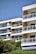 A modern apartment building with multiple balconies featuring glass railings. The structure is surrounded by lush greenery and trees, and there are blue and white awnings on some balconies. The clear blue sky complements the white and beige tones of the building.