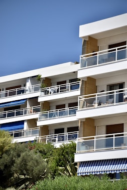 A modern apartment building with multiple balconies featuring glass railings. The structure is surrounded by lush greenery and trees, and there are blue and white awnings on some balconies. The clear blue sky complements the white and beige tones of the building.