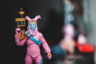 A toy figure dressed in a pink outfit resembling a bunny suit, holding a cylindrical object with a black and orange color scheme. The head has a blue mask with decorative features, and there is a blue strap across the chest. The background is blurred, focusing attention on the figure.