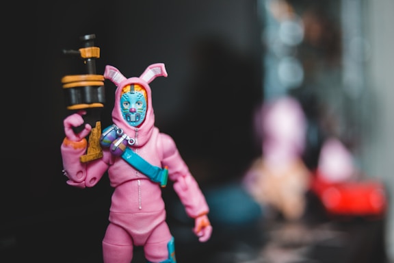 A toy figure dressed in a pink outfit resembling a bunny suit, holding a cylindrical object with a black and orange color scheme. The head has a blue mask with decorative features, and there is a blue strap across the chest. The background is blurred, focusing attention on the figure.
