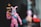 A toy figure dressed in a pink outfit resembling a bunny suit, holding a cylindrical object with a black and orange color scheme. The head has a blue mask with decorative features, and there is a blue strap across the chest. The background is blurred, focusing attention on the figure.