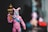 A toy figure dressed in a pink outfit resembling a bunny suit, holding a cylindrical object with a black and orange color scheme. The head has a blue mask with decorative features, and there is a blue strap across the chest. The background is blurred, focusing attention on the figure.