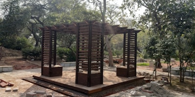 A wooden pergola stands in a garden area surrounded by lush green trees and foliage. The pergola features an open design with slatted panels and a flat roof, and there is a paved pathway nearby. Construction materials such as bricks and stones are scattered around the site, suggesting ongoing landscaping work.