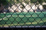 A chain-link fence is prominently featured in the foreground with a blurred view of a green sports field in the background. The field has white lines typical of a sports ground. Trees are visible, creating a natural border around the field.