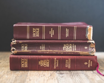 Three aged Bibles with worn covers stacked on a wooden surface against a plain dark background. The books exhibit signs of extensive use with frayed edges and faded text.