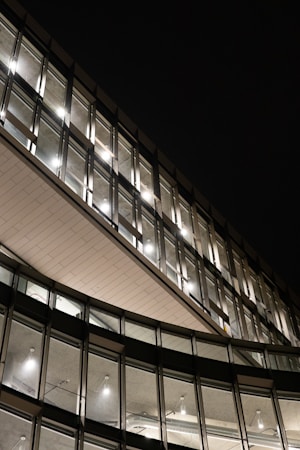 The image displays the exterior of a modern, multi-story building with large glass windows. The architectural design features geometric lines and a curved section. Bright interior lights are visible through the windows, creating a contrast against the dark night sky.