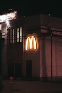 A dimly lit street scene featuring a building with an illuminated golden emblem in the shape of a large 'M' on its exterior. The structure appears to be part of a chain fast-food restaurant. The ambient lighting casts a warm glow on the surrounding surfaces, and there are various architectural details such as windows and doors visible.