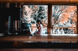 A child kneels in a prayerful pose inside a wooden structure, suggesting a moment of reflection or spirituality. The background shows blurred figures and trees with autumn-colored leaves.