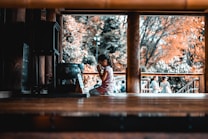 A child kneels in a prayerful pose inside a wooden structure, suggesting a moment of reflection or spirituality. The background shows blurred figures and trees with autumn-colored leaves.