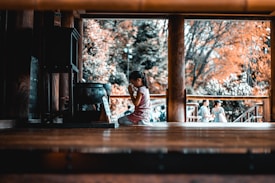 A child kneels in a prayerful pose inside a wooden structure, suggesting a moment of reflection or spirituality. The background shows blurred figures and trees with autumn-colored leaves.