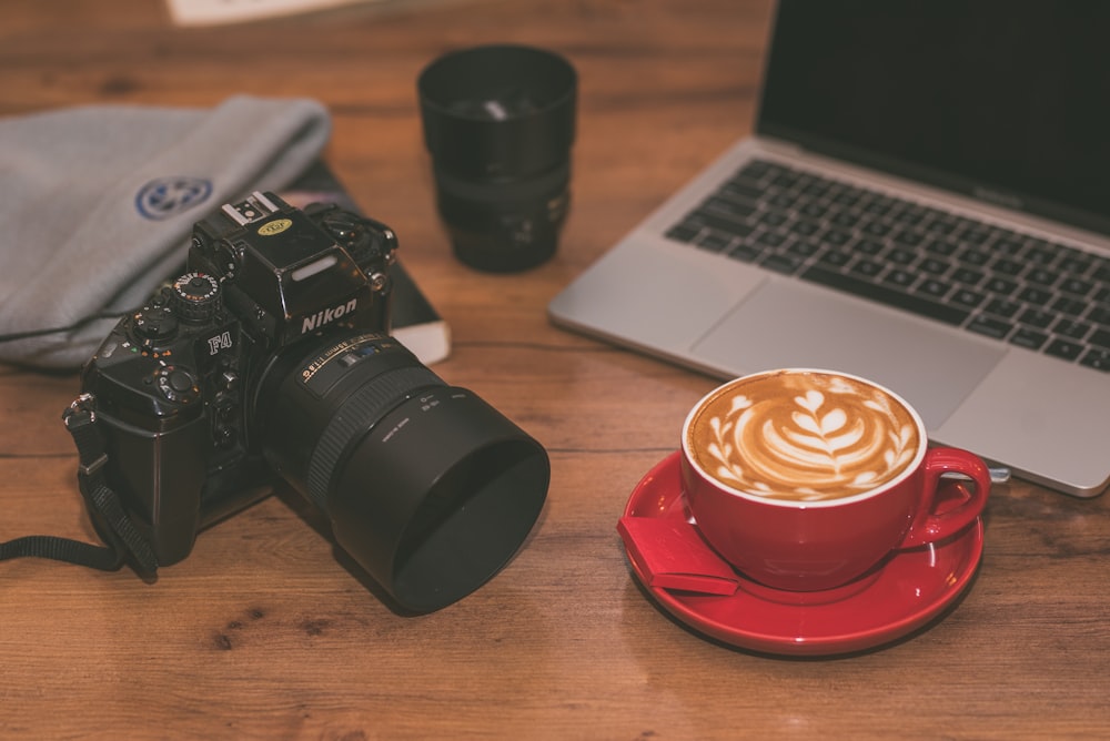 cappuccino in red ceramic mug near MacBook Pro, black camera lens, and black Nikon DSLR camera