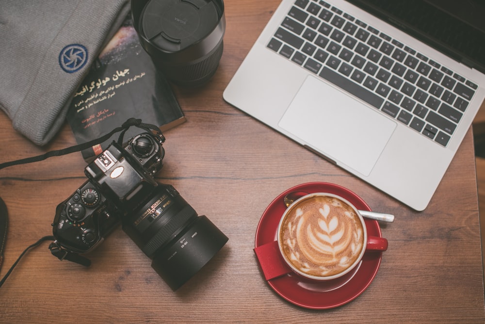 cappuccino in red ceramic mug near MacBook Pro, black camera lens, and black DSLR camera