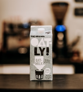 A carton of Oatly oat milk is prominently placed on a table. The carton features bold black and white text with the brand's name 'OATLY!' clearly visible, along with details about the product being vegan and free from dairy, nuts, and gluten. The background is slightly blurred, suggesting an indoor setting, possibly a kitchen, with an out-of-focus coffee maker positioned behind the carton.