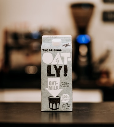 A carton of Oatly oat milk is prominently placed on a table. The carton features bold black and white text with the brand's name 'OATLY!' clearly visible, along with details about the product being vegan and free from dairy, nuts, and gluten. The background is slightly blurred, suggesting an indoor setting, possibly a kitchen, with an out-of-focus coffee maker positioned behind the carton.