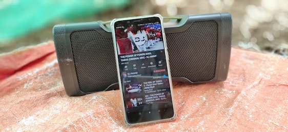 A smartphone displays a YouTube video in front of a portable speaker. The phone features a music playlist on the screen, and both the phone and speaker rest on a surface covered with a reddish-brown cloth. The background is softly blurred, emphasizing the devices.