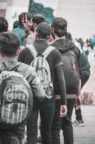 A group of young people wearing casual clothing and backpacks stands closely together. Some are facing away from the camera, and one is holding a smartphone. The setting appears to be outdoors, possibly in a public place or campus area.