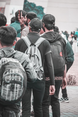 A group of young people wearing casual clothing and backpacks stands closely together. Some are facing away from the camera, and one is holding a smartphone. The setting appears to be outdoors, possibly in a public place or campus area.