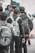 A group of young people wearing casual clothing and backpacks stands closely together. Some are facing away from the camera, and one is holding a smartphone. The setting appears to be outdoors, possibly in a public place or campus area.