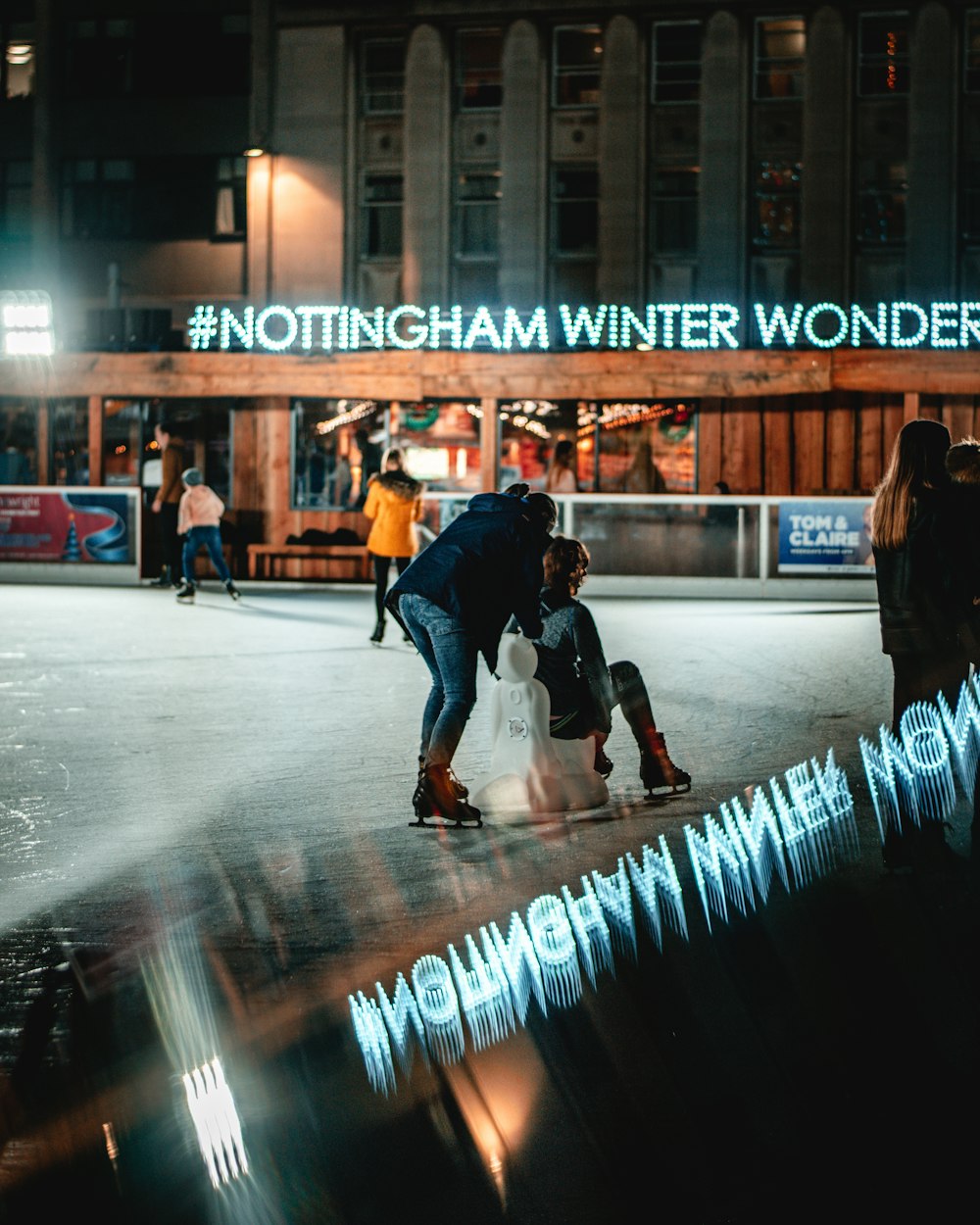 person sitting on white chair and another person standing viewing Nottingham Winter Wonderland building during night time