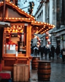 A blurred view of a festive marketplace with wooden stalls adorned with string lights. In the foreground, the focus is on a wooden kiosk, while in the background, people are walking along a street lined with grand buildings. The scene is decorated with hanging ornaments, creating a warm and festive atmosphere.