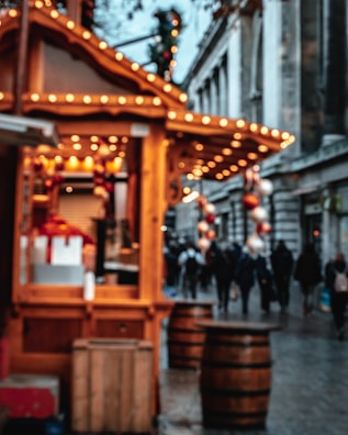 A blurred view of a festive marketplace with wooden stalls adorned with string lights. In the foreground, the focus is on a wooden kiosk, while in the background, people are walking along a street lined with grand buildings. The scene is decorated with hanging ornaments, creating a warm and festive atmosphere.