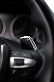 A close-up view of a car's steering wheel with a focus on the gear shift paddle. The paddle, marked with a plus sign, indicates its function for increasing gears. The interior is modern, featuring sleek design elements like metallic finish and clearly marked buttons.