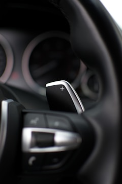 A close-up view of a car's steering wheel with a focus on the gear shift paddle. The paddle, marked with a plus sign, indicates its function for increasing gears. The interior is modern, featuring sleek design elements like metallic finish and clearly marked buttons.