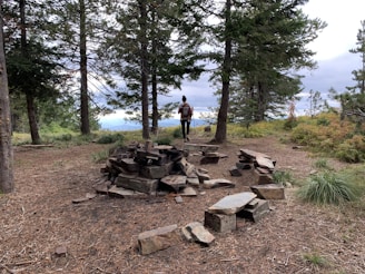 A person stands in a forest clearing, surrounded by pine trees and scattered rocks. The ground is covered with dry leaves and twigs. The sky is overcast, adding a sense of calm to the natural setting.