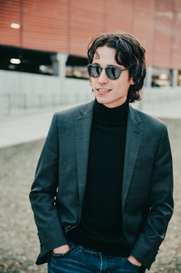man wearing gray notched lapel suit jacket and sunglasses standing on field smiling