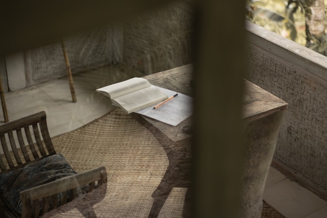 An open book lies on a rustic wooden desk, accompanied by a pencil resting on some papers. Next to the desk, a wooden chair with slatted back and a cushion is positioned on a textured woven rug. The space is enclosed by walls with a woven pattern and a window providing natural light.